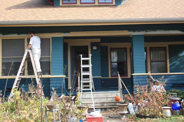 A painter on a ladder painting the porch of a blue house